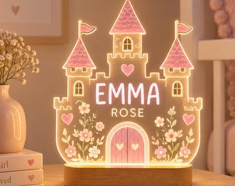 May include: A light-up acrylic castle night light with a wooden base. The castle features pink roofs, floral accents, and heart shapes. The text EMMA ROSE is printed in the center. The design includes three towers with flags and a central arched door.