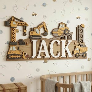 May include: Wooden construction-themed name sign with the name "JACK" in white letters. The sign features various construction vehicles in yellow, gray, and brown, including a crane, excavator, and dump truck. The sign is mounted on a light brown wooden base.