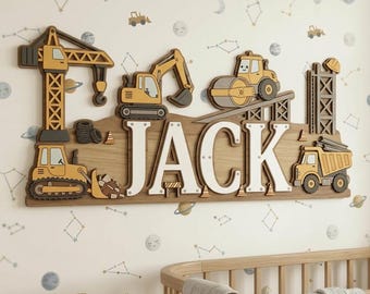 May include: Wooden construction-themed name sign with the name "JACK" in white letters. The sign features various construction vehicles in yellow, gray, and brown, including a crane, excavator, and dump truck. The sign is mounted on a light brown wooden base.
