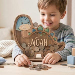 May include: Wooden dinosaur-shaped piggy bank with the name "NOAH" on it. The dinosaur is light brown with blue and green accents. The bank is filled with coins, and a child is placing more coins into the slot.