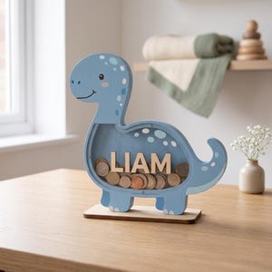 May include: A light blue wooden dinosaur-shaped coin bank with the name "LIAM" in wooden letters. The bank is filled with coins and sits on a wooden base. The dinosaur has white spots and a smiling face.