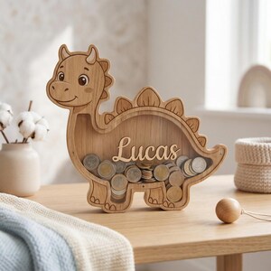 May include: Wooden dinosaur-shaped coin bank filled with coins. The bank is light brown with the name "Lucas" in script. The dinosaur has a friendly face and a row of spikes along its back. The bank is sitting on a wooden table.