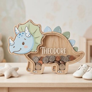 May include: A wooden triceratops-shaped coin bank with the name "Theodore" in white script. The dinosaur is light brown with blue and green accents. The bank is filled with coins and sits on a light-colored surface.