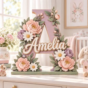 Könnte beinhalten: Ein großer, hellvioletter Holzbuchstabe A mit dem Namen Amelia in weißer Schreibschrift in der Mitte. Der Buchstabe ist mit verschiedenen künstlichen Blumen verziert, darunter rosa Pfingstrosen, weiße Gänseblümchen und grüne Blätter.
