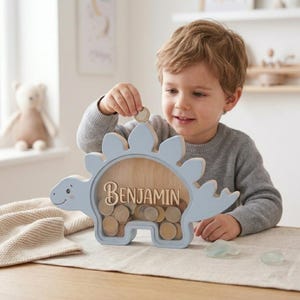 May include: A light blue dinosaur-shaped wooden piggy bank with the name "BENJAMIN" in wooden letters. The piggy bank is filled with coins. A young child is putting a coin into the slot.