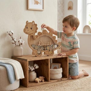 May include: A wooden dinosaur-shaped piggy bank with the name "Lucas" engraved on it. The bank is filled with coins and a child is adding more. The bank is sitting on a wooden shelf with decorative items.