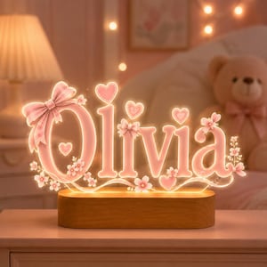 Op de afbeelding: Een gepersonaliseerd lichtbord met de naam Olivia in zachtroze letters. Het ontwerp bevat decoratieve elementen zoals een grote strik op de eerste letter, kleine hartvormen en delicate kersenbloesems, gemonteerd op een houten voet.