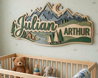May include: Wooden mountain-themed name sign with the names "Julian" and "Arthur" in green script. The sign features layered mountains, trees, a river, and a cabin, with a moon and stars. The sign is mounted above a wooden crib.