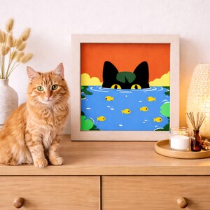 Pode incluir: Uma impressão artística emoldurada retrata um gato preto espreitando de um lago com peixes amarelos. A obra de arte é definida contra um céu laranja e água azul. Um gato malhado laranja está sentado ao lado da obra de arte emoldurada.