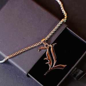 May include: A silver-toned metal necklace featuring a pendant shaped like the letter L in a gothic-style font. The pendant is suspended from a box chain, and the entire piece is displayed resting inside a small, open black rectangular jewelry box.