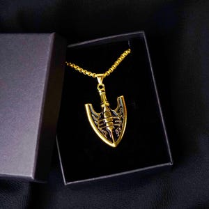 May include: A gold-coloured pendant necklace with a distinctive arrowhead design, displayed in a black gift box. The pendant has intricate black detailing and hangs from a matching gold chain. A fashionable accessory.