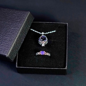 Op de afbeelding: Een sieradenset bestaande uit een ketting en een ring in een zwarte doos. De ketting heeft een hanger van donker metaal met een grote, ovaalvormige paarse edelsteen. De ring heeft een bijpassende band met een ronde paarse steen en bladvormen.