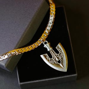 Puede incluir: Un collar de metal plateado con una cadena de eslabones y un colgante en forma de punta de flecha con intrincados calados y un pequeño orificio en forma de corazón en la base. La joya se presenta dentro de una caja de regalo gris oscuro.