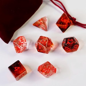 May include: A set of seven polyhedral dice with red numbering, displayed on a white surface. The dice are transparent with a red and orange marbled effect. A burgundy velvet dice bag is in the background.