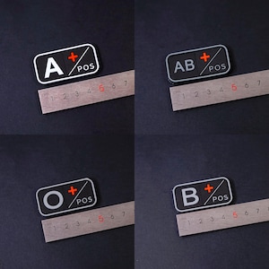 Puede incluir: Un conjunto de cuatro parches rectangulares de grupo sanguíneo con las letras A, AB, O y B en blanco, cada uno con un signo más rojo y el texto POS. Una regla metálica situada bajo cada parche muestra una anchura de unos 5 cm.