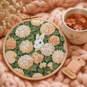 May include: A circular wooden embroidery hoop containing a cross-stitch pattern of a white rabbit nestled among clusters of peach and cream-colored hydrangea flowers with dark green leaves, accompanied by a wooden thread spool and a ceramic mug.