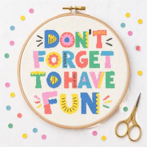 Könnte beinhalten: Ein runder Stickrahmen aus Holz mit einer Kreuzsticharbeit auf weißem Stoff. Das Design zeigt den bunten Schriftzug DON'T FORGET TO HAVE FUN in verschiedenen Schriftarten und Mustern mit blauem, rosa, grünem, gelbem und rotem Garn.
