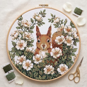 May include: A cross-stitch embroidery of a squirrel surrounded by white and cream-colored flowers and green foliage, set in a wooden hoop. The embroidery is accompanied by green thread, rose petals, and gold scissors.