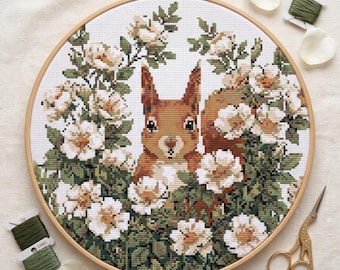 May include: A cross-stitch embroidery of a squirrel surrounded by white and cream-colored flowers and green foliage, set in a wooden hoop. The embroidery is accompanied by green thread, rose petals, and gold scissors.
