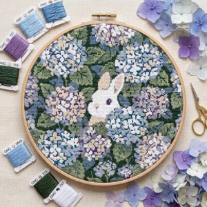 Könnte beinhalten: Kreuzstich-Stickerei in einem Holzrahmen, die ein weißes Kaninchen inmitten blauer, violetter und grüner Hortensien zeigt. Das Bild enthält Garnrollen in verschiedenen Farbtönen, eine goldene Schere und Hortensienblüten. Ein detailliertes Handarbeitsprojekt.
