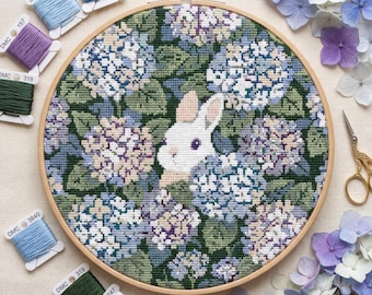 May include: A cross-stitch embroidery hoop featuring a white rabbit peeking out from a field of blue, purple, and green hydrangeas. The embroidery is set in a wooden hoop, with spools of thread in various shades of blue, green, and purple. A pair of gold scissors and hydrangea blooms are also present.