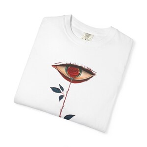 以下が含まれることがあります： 白いTシャツ。瞳孔の代わりに赤いバラが描かれた目のデザイン。目は茶色とベージュの色調で詳細に描かれ、バラは茎と葉につながっています。Tシャツは折りたたまれています。