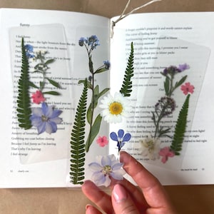 Könnte beinhalten: Drei rechteckige, transparente Lesezeichen mit gepressten botanischen Elementen, darunter grüne Farnwedel, weiße Gänseblümchen, blaue Rittersporne und kleine rosa Blüten. Ein Lesezeichen ist mit einer dünnen, hellen Schnur versehen.