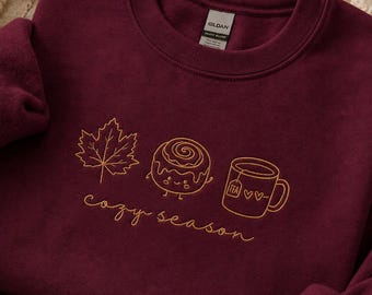 Könnte beinhalten: Ein kastanienbraunes Langarm-Sweatshirt mit goldener Stickerei, die ein Ahornblatt, eine lächelnde Zimtschnecke und eine Teetasse mit der Aufschrift "tea" und zwei Herzen zeigt. Unter den Symbolen ist "cozy season" in goldener Schreibschrift gestickt.
