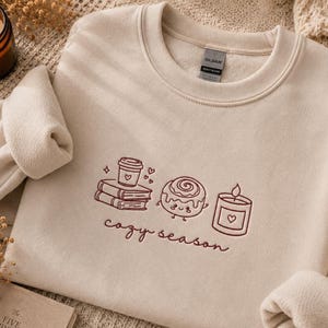 Könnte beinhalten: Ein beigefarbenes Sweatshirt mit Rundhalsausschnitt und dunkelbrauner Stickerei, die einen Bücherstapel mit Kaffeetasse, eine lächelnde Zimtschnecke und eine brennende Kerze zeigt. Der Schriftzug "cozy season" ist in Schreibschrift darunter gestickt.