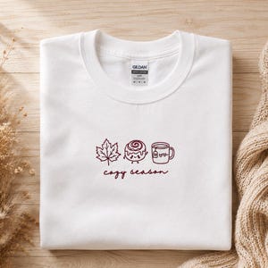 K&ouml;nnte beinhalten: Ein wei&szlig;es T-Shirt mit Rundhalsausschnitt und einer dunkelroten Stickerei auf der Brust. Das Motiv zeigt ein Ahornblatt, eine l&auml;chelnde Zimtschnecke und eine Tasse mit Teebeutel, erg&auml;nzt durch den Schriftzug "cozy season" in Schreibschrift.