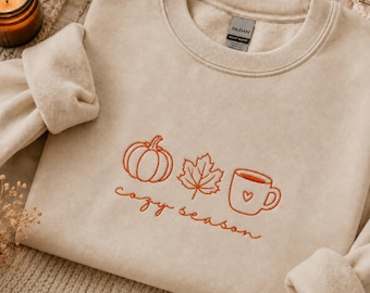 Könnte beinhalten: Ein sandfarbenes Sweatshirt mit Rundhalsausschnitt und orangefarbener Stickerei eines Kürbisses, eines Ahornblatts und einer Tasse mit einem kleinen Herz. Der Text "cozy season" ist in orangefarbener Schreibschrift unter den Symbolen gestickt.