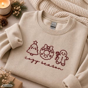 Könnte beinhalten: Ein beigefarbenes Sweatshirt mit Rundhalsausschnitt und dunkelroter Stickerei, die einen lächelnden Weihnachtsbaum, einen Kranz mit Schleife und ein Lebkuchenmännchen zeigt. Der Text "cozy season" ist in Schreibschrift darunter gestickt.