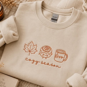 Könnte beinhalten: Ein sandfarbenes Sweatshirt mit Rundhalsausschnitt und rostfarbener Stickerei eines Ahornblatts, einer lächelnden Zimtschnecke und einer Teetasse mit zwei Herzen. Der Text "cozy season" ist in Schreibschrift unter den Motiven gestickt.
