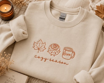 Könnte beinhalten: Ein sandfarbenes Sweatshirt mit Rundhalsausschnitt und rostfarbener Stickerei eines Ahornblatts, einer lächelnden Zimtschnecke und einer Teetasse mit zwei Herzen. Der Text "cozy season" ist in Schreibschrift unter den Motiven gestickt.