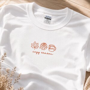 K&ouml;nnte beinhalten: Ein wei&szlig;es T-Shirt mit Rundhalsausschnitt und einer Stickerei in orangefarbenem Garn. Das Motiv zeigt ein Ahornblatt, eine Zimtschnecke mit Gesicht und eine Tasse mit zwei Herzen, gefolgt von dem kursiven Text "cozy season" unter den Symbolen.