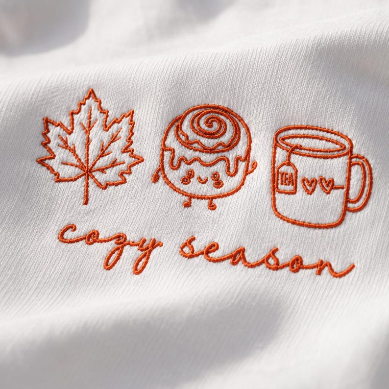 K&ouml;nnte beinhalten: Ein wei&szlig;er Stoff mit orangefarbener Stickerei eines Ahornblatts, einer l&auml;chelnden Zimtschnecke und einer Tasse mit Teebeutel und zwei Herzen. Unter den Symbolen ist der Text "cozy season" in orangefarbener Schreibschrift auf den Stoff gestickt.