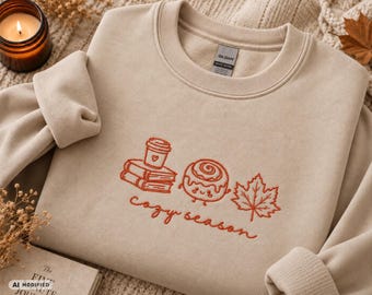 Könnte beinhalten: Ein sandfarbenes Sweatshirt mit Rundhalsausschnitt und rostfarbener Stickerei, die einen Bücherstapel mit einer Kaffeetasse, eine lächelnde Zimtschnecke und ein Ahornblatt zeigt, sowie den kursiven Schriftzug "cozy season" darunter.