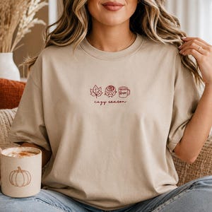 K&ouml;nnte beinhalten: Ein beiges Kurzarm-T-Shirt mit einer kleinen Stickerei auf der Brust, die ein Ahornblatt, eine Zimtschnecke und eine Tasse zeigt, darunter der Text "cozy season". Dazu geh&ouml;rt eine gesprenkelte Tasse mit einer K&uuml;rbisgrafik.