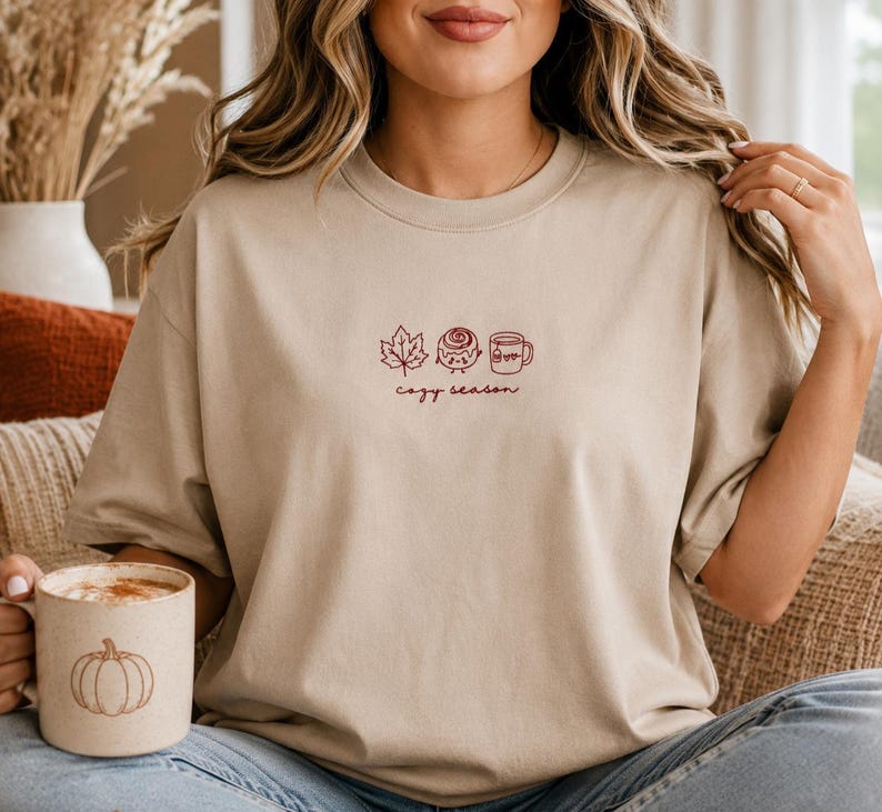 K&ouml;nnte beinhalten: Ein beiges Kurzarm-T-Shirt mit einer kleinen Stickerei auf der Brust, die ein Ahornblatt, eine Zimtschnecke und eine Tasse zeigt, darunter der Text "cozy season". Dazu geh&ouml;rt eine gesprenkelte Tasse mit einer K&uuml;rbisgrafik.