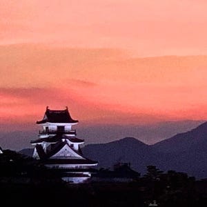 以下が含まれることがあります： 鮮やかな夕焼けを背景にした日本の城。 多層構造は、暗い屋根と白い壁が特徴で、紫色の山々とピンクとオレンジの色合いのグラデーションの空を背景にしています。