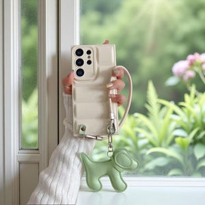 May include: Cream-colored phone case with a quilted design and a matching wrist strap. A light green dog-shaped charm hangs from the case. The phone case is held in front of a window with a green background.