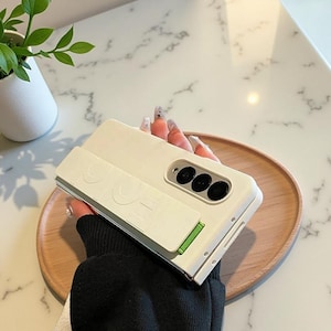 May include: A white phone case with a built-in stand and a green accent. The case has a rectangular design with the word "LOVE" embossed on the front. The case has three black camera lenses.