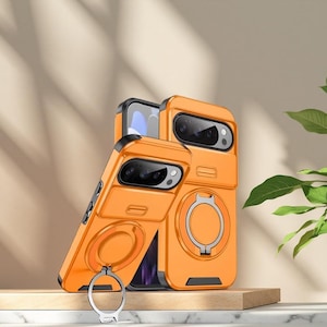 May include: Orange phone cases with a built-in ring holder and camera protection. The cases are shown in a studio setting, with a wooden block and a plant in the background. The ring holder is silver.