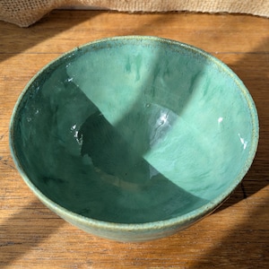 May include: A teal ceramic bowl with a glossy finish. The bowl has a rounded shape and a slightly textured surface. The interior reflects light, and the rim has a darker shade of teal. The bowl is on a wooden surface. The bowl is likely handmade.
