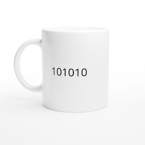 May include: White ceramic coffee mug with a curved handle. The mug features the black binary code "101010" printed on the side. The mug is set against a white background.