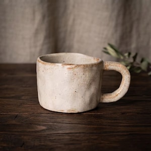 Puede incluir: Una taza de cerámica con una superficie texturizada en color blanco roto y un asa gruesa y redondeada. La taza presenta bordes irregulares formados a mano y sutiles motas marrones a lo largo del borde y el asa, con un acabado mate.
