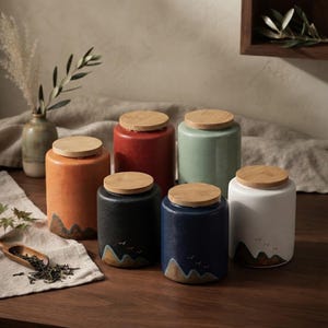Puede incluir: Un conjunto de seis botes de almacenamiento cilíndricos de cerámica con tapas planas de bambú. Los botes presentan varios colores sólidos, incluyendo naranja, rojo, verde claro, negro, azul oscuro y blanco, con motivos de montañas y aves.
