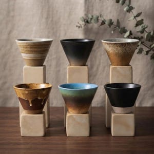 May include: Six ceramic cups with varied glazes, including tan, black, speckled brown, drip-patterned brown, blue-to-brown gradient, and black-and-white, each resting on a square beige block, arranged in two rows of three on a wooden surface.