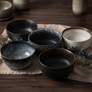 May include: A collection of five ceramic bowls featuring varied finishes, including solid matte black, speckled dark gray, and a cream-colored bowl with a brown rim. The bowls are arranged in a cluster, showcasing different textures and glaze patterns.