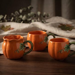 Peut inclure: Trois tasses en céramique orange en forme de citrouille, chacune dotée d'une anse verte et d'un petit motif de feuille et de vigne. Les tasses ont une finition brillante et des corps arrondis, l'une présentant un bord festonné.